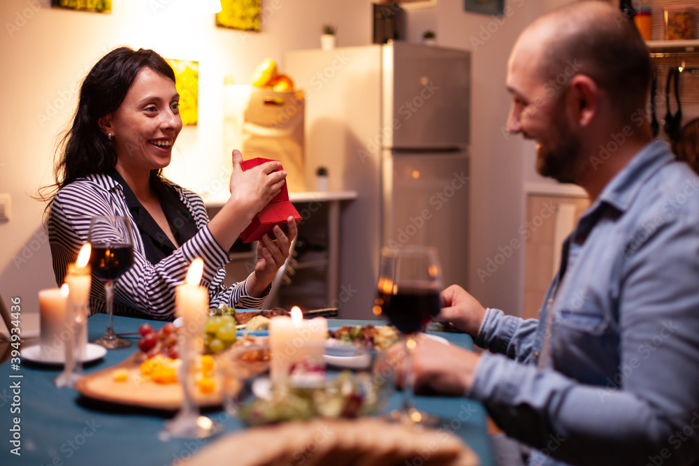 Woman holding anniversary gift from husband during dinner. Happy cheerful couple dining together at home, enjoying the meal celebrating their anniversary. Woman holding anniversary gift from husband during dinner. Happy cheerful couple dining together at home, enjoying the meal celebrating their anniversary.