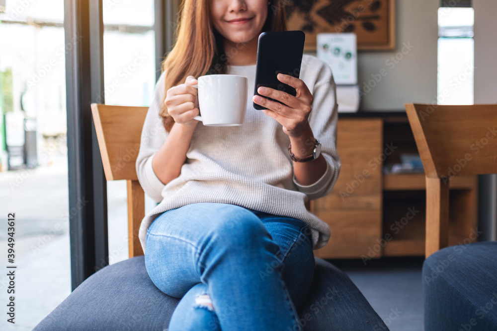 Obraz premium Closeup image of a beautiful asian woman holding and using mobile phone while drinking coffee in cafe