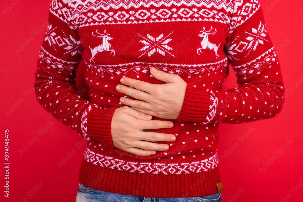 Bellyache concept. Close up photo of men in red winter sweater with deers holding his stomach isolated on red background Bellyache concept. Close up photo of men in red winter sweater with deers holding his stomach isolated on red background