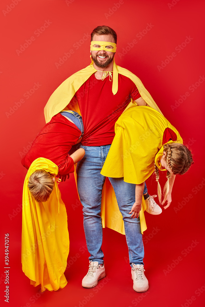 positive woman try to calm down playful children, holding them in two hands, wearing superhero cloak and mask positive woman try to calm down playful children, holding them in two hands, wearing superhero cloak and mask