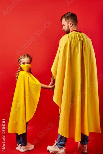 friendly man and kid girl in yellow cloaks, pretend to be superheroes. male and child in mask stand holding hands together, rear view, girl looks at camera
