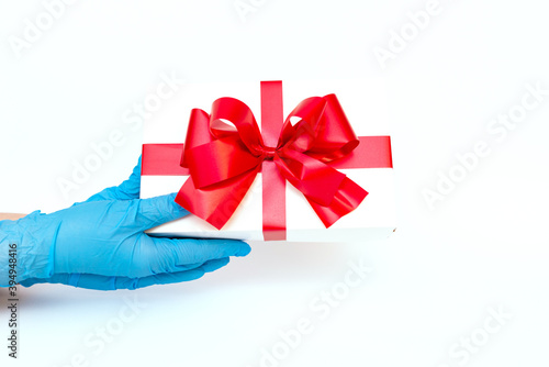 Holiday gift.Hands wearing meidical gloves holding gift box with red ribbon.white background.COVID-19 prevention. photo