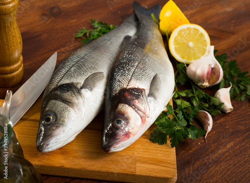 Two whole fresh European basses lying on cutting board with lemon, greens and seasonings on wooden surface photo