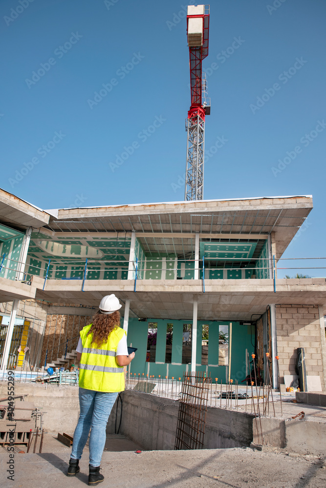 Architect supervising construction site, young female professional wearing a hardhat and neon safety jacket while holding an electronic device comparing construction stage and progress with blueprints
