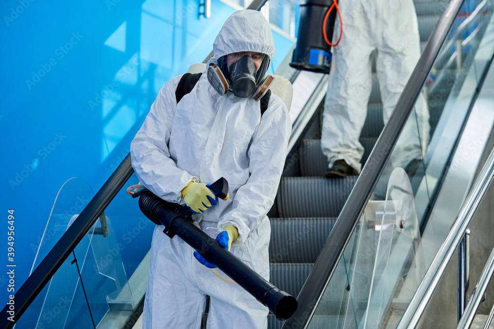 male disinfector cleaning the area in building, disinfect escalator, wearing protective white suit, getting rid of coronavirus infections around