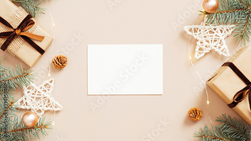 Christmas flat lay composition with empty paper card mockup, gift boxes, stars decoration, fir tree branches on beige background. Cozy home table, hygge style.
