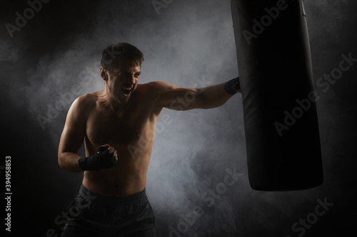 Aggressive boxer in black boxing wraps punching in boxing bag on dark background with smoke © producer