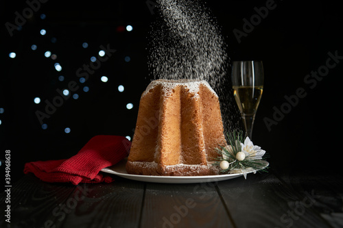 Celebrating Christmas with a snowfall of powdered sugar that whitens a pandoro, a typical dessert of the Christmas tradition, with a glass of sparkling wine and a bokeh effect on a dark background.