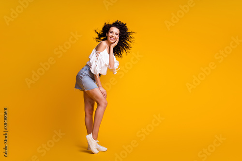 Full size profile photo of positive funky curly brunette woman standing wearing white top blue skirt sneakers isolated on yellow background