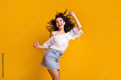 Photo of young cheerful cute curly brunette woman dancing wearing white top blue skirt isolated on vivid yellow colored background