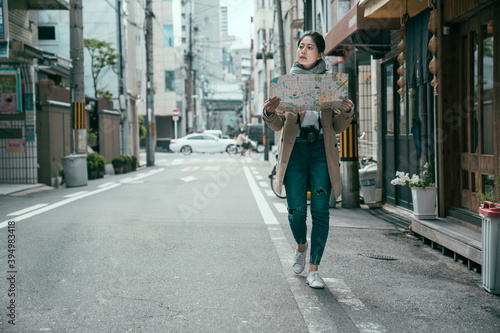 full length asian teenage woman looking away for destination while holding paper map for taking route around osaka city. female traveler with vintage camera spending time for trip walking on road