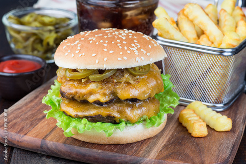 Double cheese burger with two beef patty, cheddar cheese, lettuce, caramelized onion and jalapeno slices, served with french fries and soda, on wooden board, horizontal