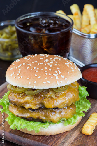 Double cheese burger with two beef patty, cheddar cheese, lettuce, caramelized onion and jalapeno slices, served with french fries and soda, on wooden board, vertical, closeup