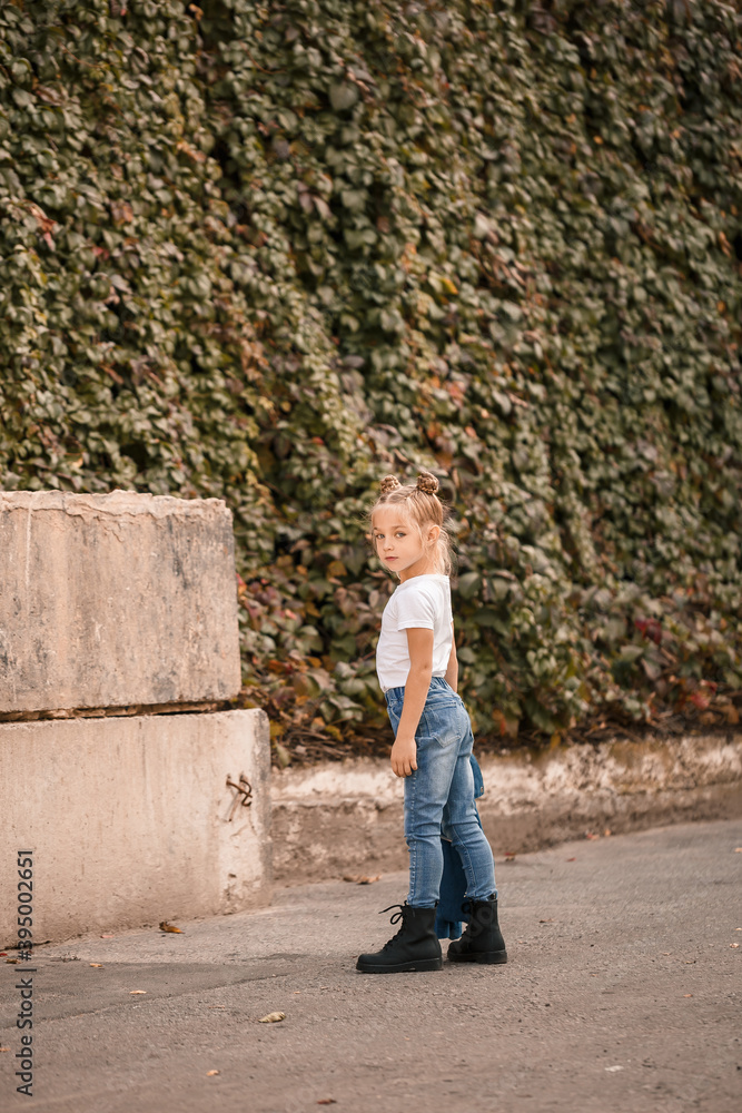 Stylish blonde little girl in jeans and a white T-shirt walks down the street. Girl 7 years old small model, beautiful child