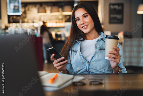 Close-up. Beautiful smiling young business woman sits in front of laptop, holds cup of coffee, uses smartphone for work