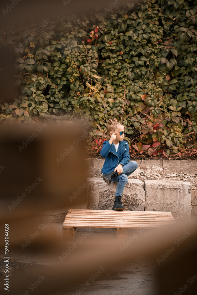 A cute little girl is sitting in sunglasses on a stone fence. Stylish child in a blue jacket. Casual style, fashion for children, fashionable suit, happy childhood A cute little girl is sitting in sunglasses on a stone fence. Stylish child in a blue jacket. Casual style, fashion for children, fashionable suit, happy childhood