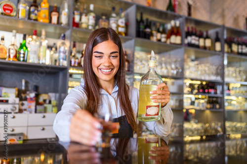 Beautiful female bartender with protective face maskholding shot glass with alcohol during coronavirus pandemic, shelves full of bottles with alcohol on the background