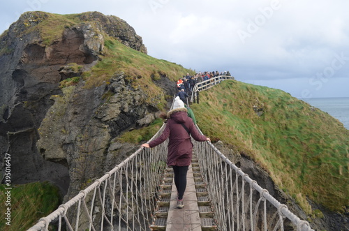 Woman crossing the Carrick-a-Rede Rope Bridge in Ireland while people look at her photo