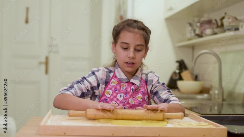 Little child girl in the kitchen dressed in apron and scarf like chef. Girl roll dough with rolling pin, smiles happy. Concept of: nutrition, cooking school, education. Little child girl in the kitchen dressed in apron and scarf like chef. Girl roll dough with rolling pin, smiles happy. Concept of: nutrition, cooking school, education.