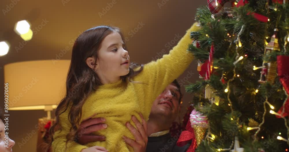 Close up of Caucasian happy parents help cute little daughter to decorate xmas tree on New Year's Eve at home. Family decorating christmas tree on holidays. Xmas decorations. New Year's eve Close up of Caucasian happy parents help cute little daughter to decorate xmas tree on New Year's Eve at home. Family decorating christmas tree on holidays. Xmas decorations. New Year's eve