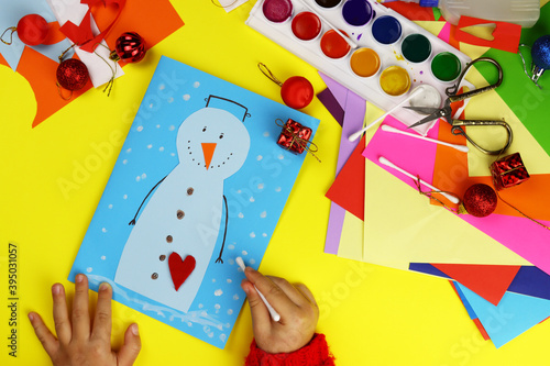 The child makes a Christmas card on the table of the house, applique snowman