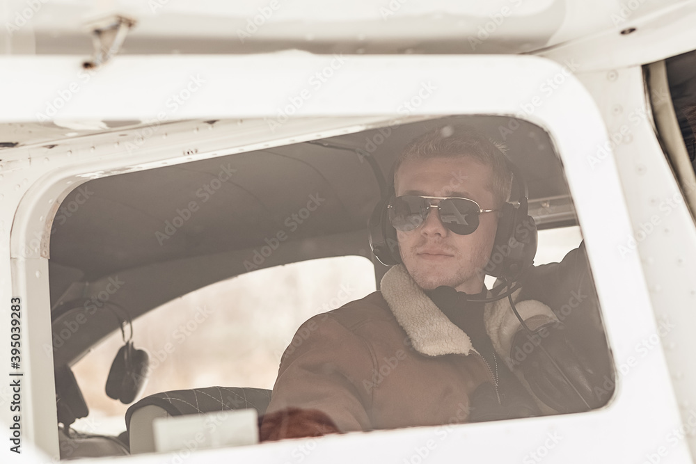 A young, handsome male pilot in winter clothes, glasses and headphones, sitting in the cockpit of a small pleasure plane. the view through the glass.