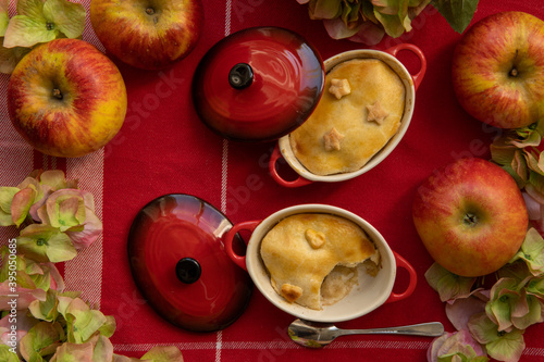 Little apple pies in pots with bright red lids on a red table cloth, decorated with apples and flowers.