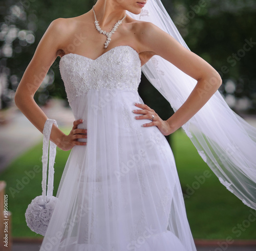 Young beautiful bride in a white wedding dress and long veil posing with a bridal stylish round purse on the green background. Wedding photography and concept.