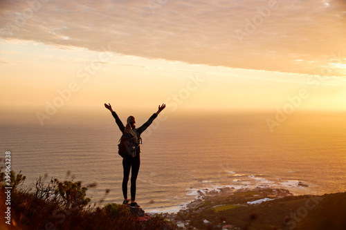 Rear View Of Active Young Woman Hiking Along Coastal Path At Sunset With Arms Outstretched