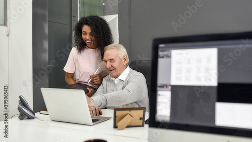 Aged man, senior intern looking at laptop while showing results to his young colleague, Friendly female worker training, mentoring new employee, preparing for work