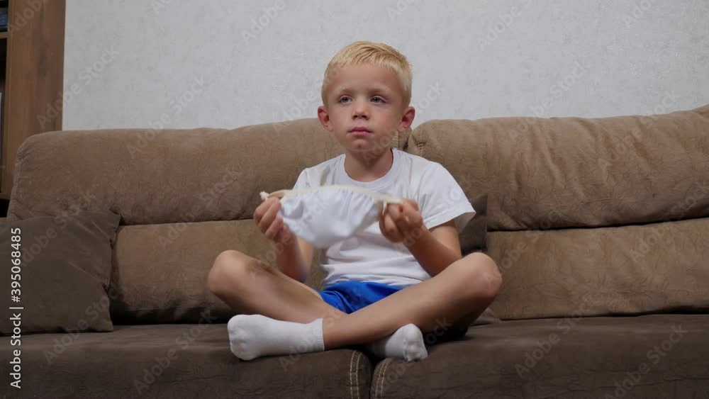 Portrait of a little boy at home on the sofa in a protective medical mask, he takes off the mask from his face. An outbreak of coronavirus. Portrait of a little boy at home on the sofa in a protective medical mask, he takes off the mask from his face. An outbreak of coronavirus.