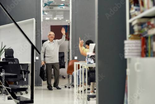 Full length shot of cheerful aged man, senior intern saying goodbye to his young colleagues while leaving office after first day at work photo