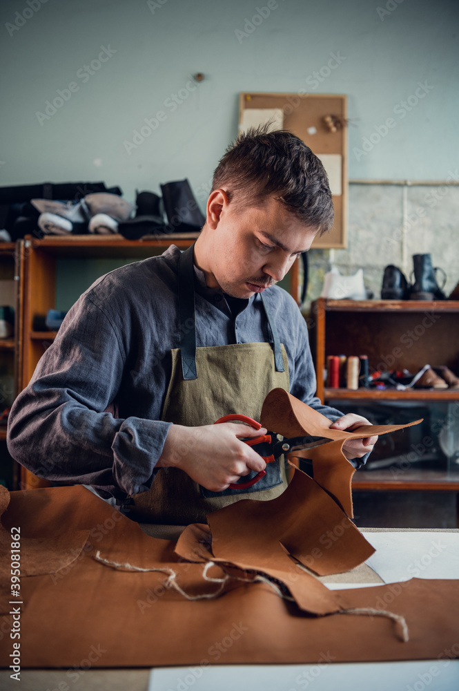 Fototapeta premium A young tanner in his workshop at the table cuts out leather elements of shoes.