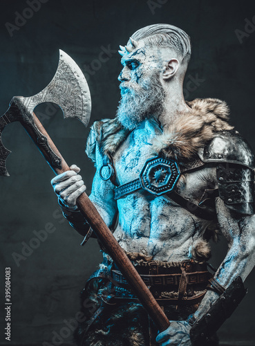 Naked and pale skinned god with horns and blue eyes holding two handed axe and looking at it in dark background.