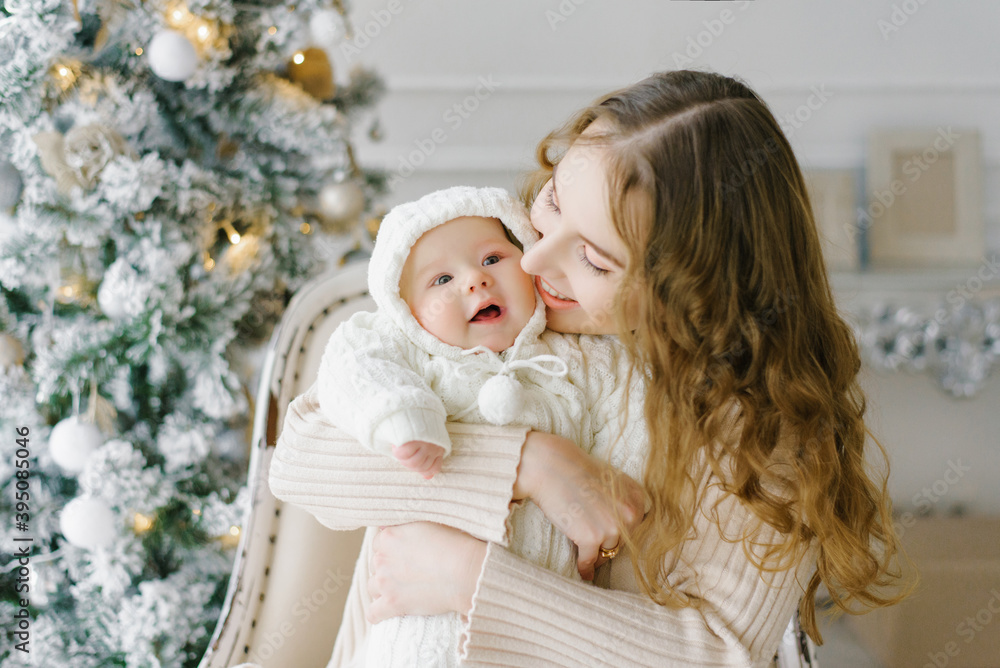 A beautiful mother holds a small child in her arms against the background of a Christmas tree. The woman looks at the child and smiles. A beautiful mother holds a small child in her arms against the background of a Christmas tree. The woman looks at the child and smiles.