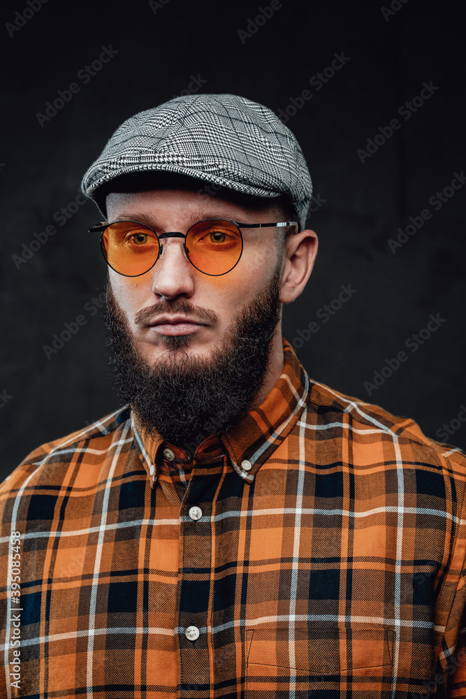 Manly and bearded hipster guy dressed in shirt with cap and weared with sunglasses in dark background.