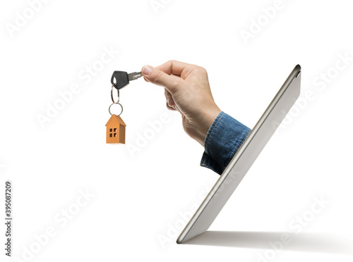 Key with paper house in human hand stick out of a digital tablet screen. Concept of modern technologies in real estate industry.