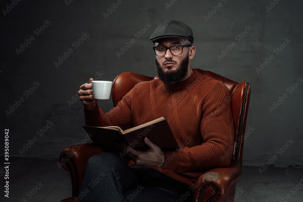 Sitting on leather armchair in dark background clever and manly guy with beard and glasses weared in sweater with cap.