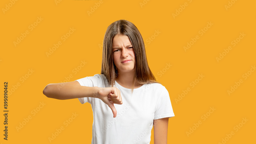 Disappointed child. Dislike gesture. Bad idea. Refusal signal. Disgusted skeptic girl showing thumb down sign isolated on orange copy space background. Disappointed child. Dislike gesture. Bad idea. Refusal signal. Disgusted skeptic girl showing thumb down sign isolated on orange copy space background.