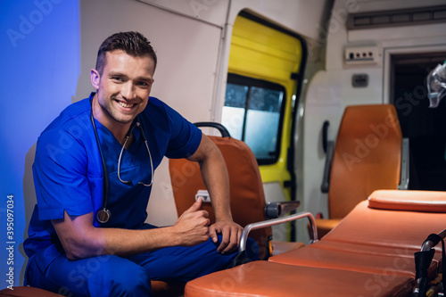 A portrait of a smiling young paramedic in a blue uniform sitting in the ambulance car and showing thumbs up.