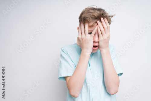 A mid shot of a frightened caucasian boy on a white background. He covered his face with his hands and now he's peeking out of his fingers