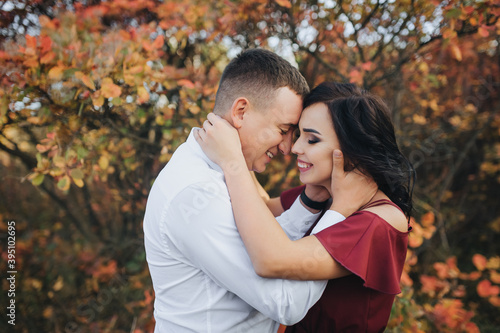 A stylish respectable guy in a white shirt and trousers and a beautiful girl in an expensive long dress stand and laugh, hugging against the background of red, yellow bushes with leaves.