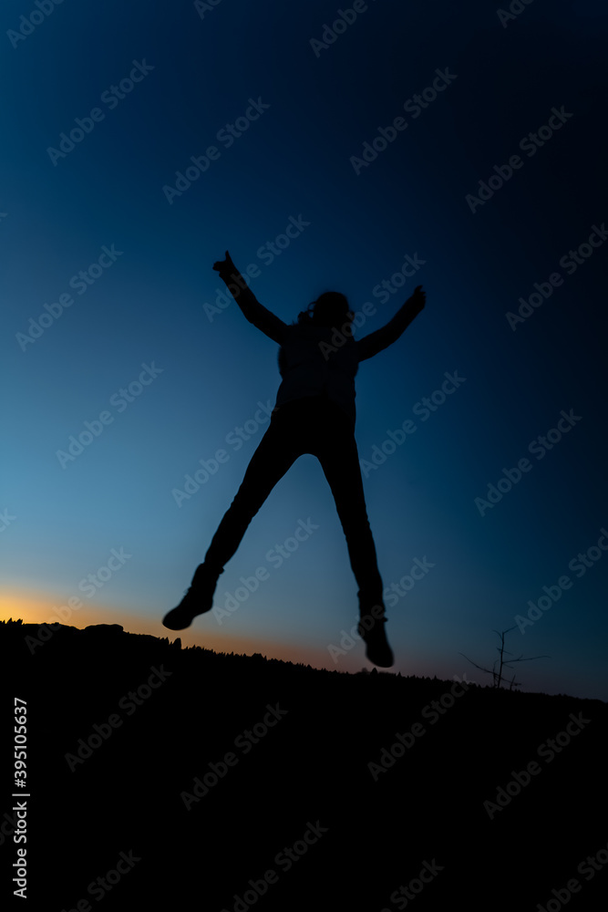 Silhouette of a girl in a jump on a background of sunset and a dark blue sky. The concept of happy people. Woman with arms outstretched jumping up