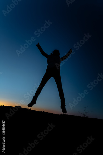 Silhouette of a girl in a jump on a background of sunset and a dark blue sky. The concept of happy people. Woman with arms outstretched jumping up