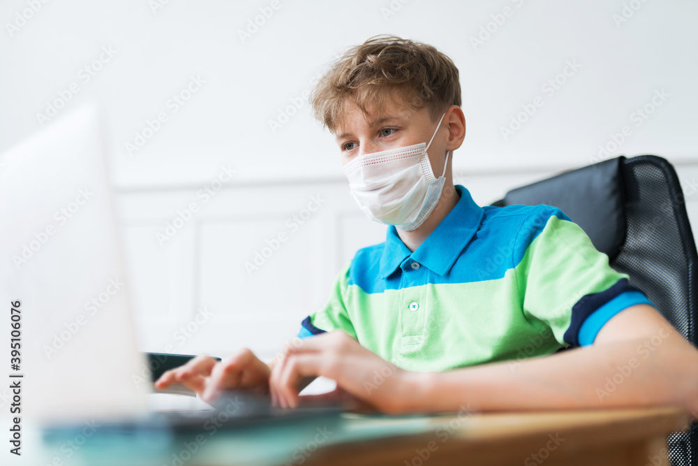 Blonde American boy in a disposable face mask is actively participating in his online classes during the lockdown. He's typing the text on his laptop and listening to the teacher Blonde American boy in a disposable face mask is actively participating in his online classes during the lockdown. He's typing the text on his laptop and listening to the teacher