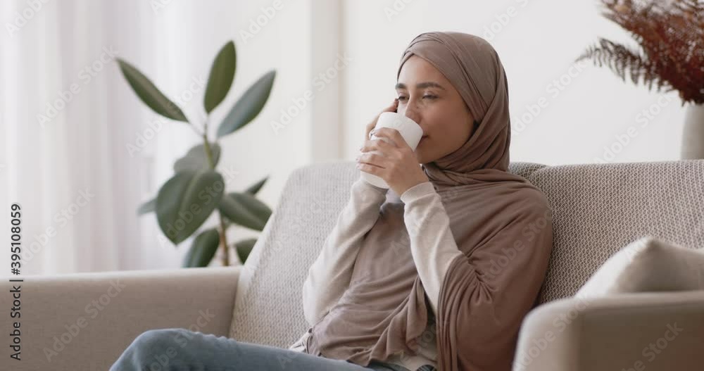 Young muslim woman talking on cellphone and enjoying hot tea, sitting on couch at home, slow motion Young muslim woman talking on cellphone and enjoying hot tea, sitting on couch at home, slow motion