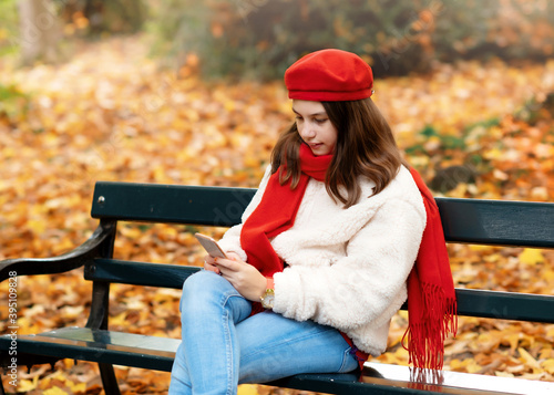 girl in red hat and scarf looking at phone and sitting on the bench in autumn park