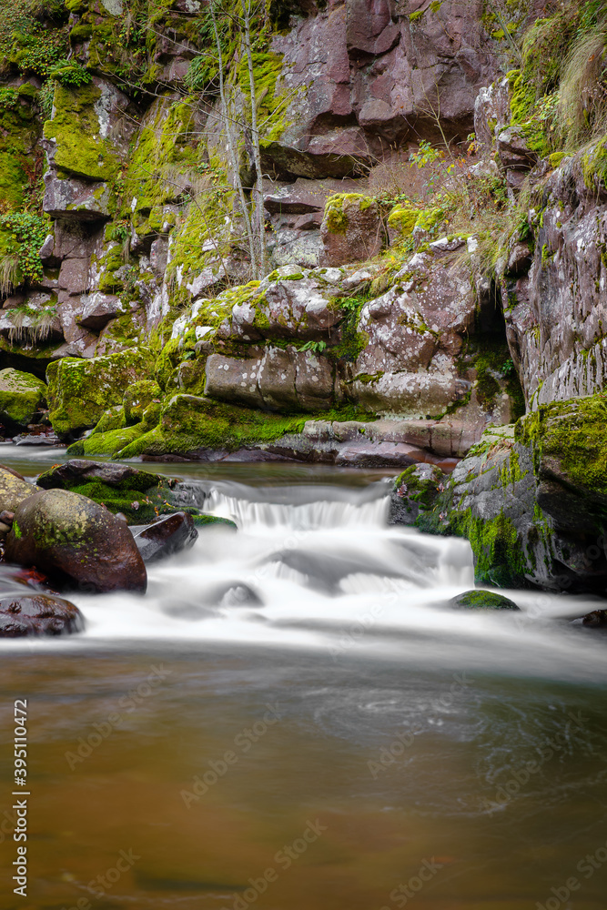 Fototapeta premium Small cascade on a mountain river flowing through the narrow, red rock canyon with rocks covered by green moss and cliffs with autumn colored trees