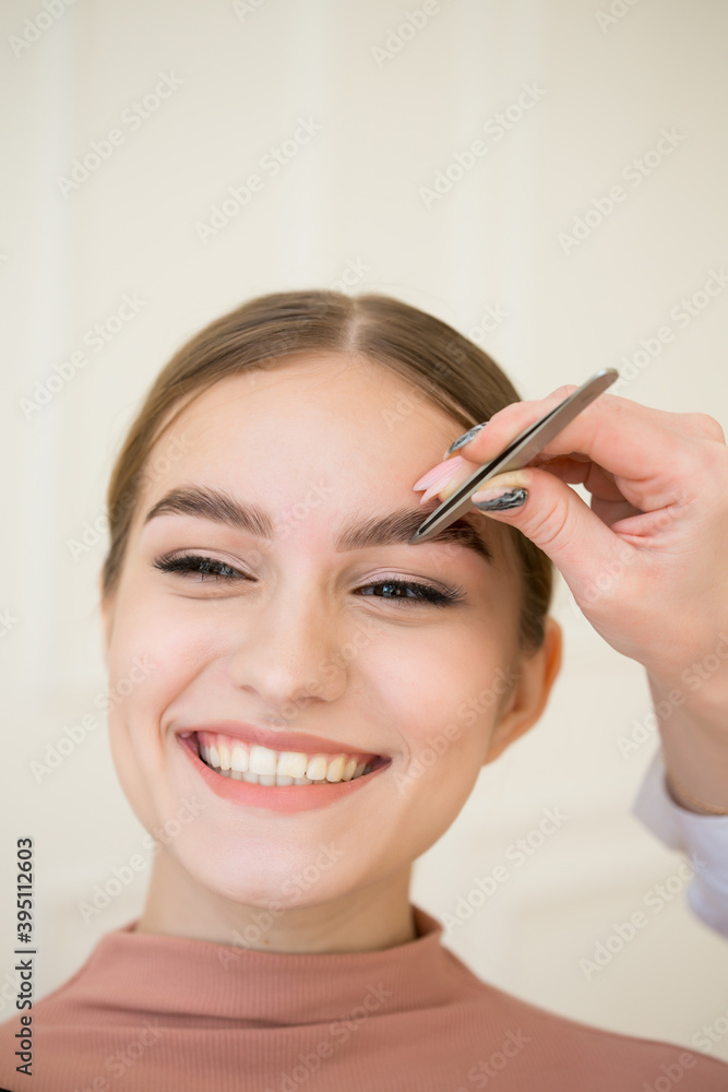 Beautiful happy caucasian girl smiling while shaping eyebrows with tweezers. Eyebrow care procedure.Cosmetology and makeup concept