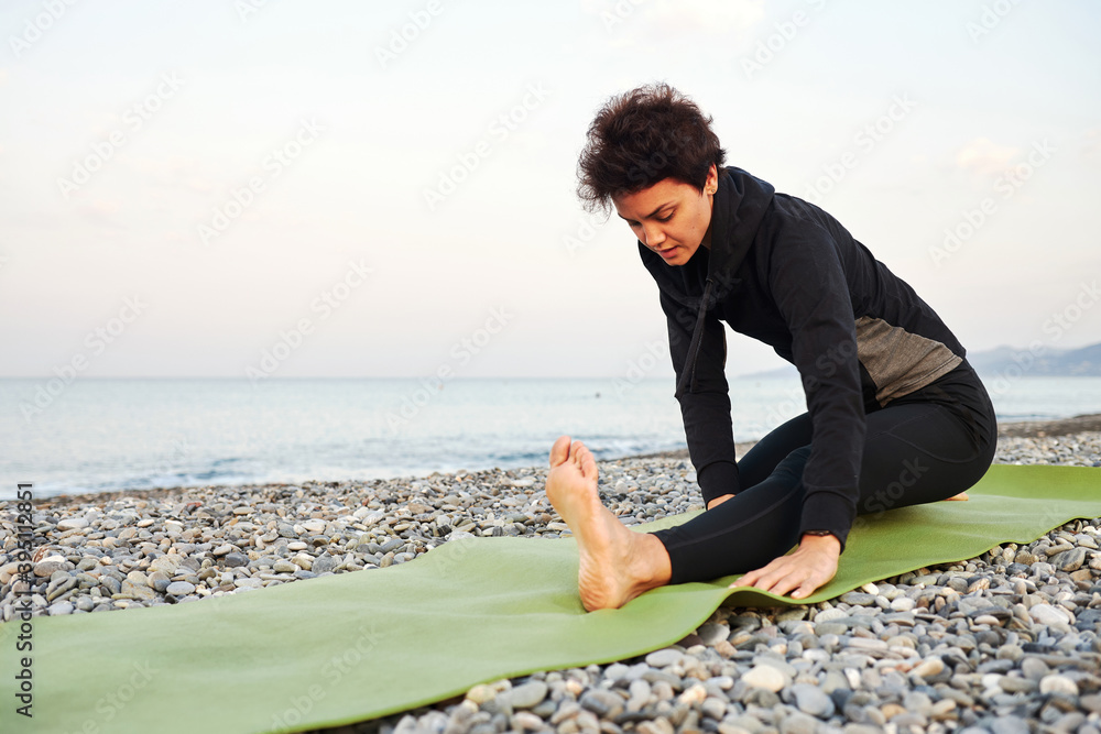Young sporty woman with short hair in black sportswear practices yoga, stretching and meditation on mat by the sea, morning exercises on nature background, relax breath